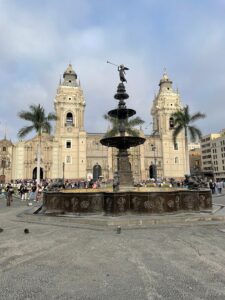 Fuente de bronce de la Plaza de Armas de Lima con la Catedral de Lima al fondo y cielo nublado.